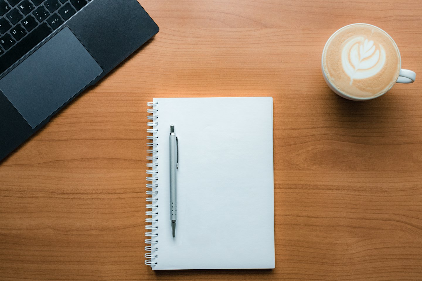 Laptop, notitieboekje, pen en koffiekopje op een houten bureau.