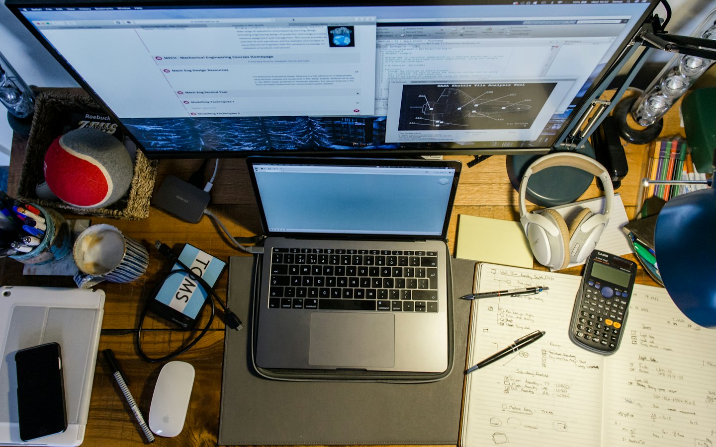Laptop en externe monitor op een rommelig houten bureau.