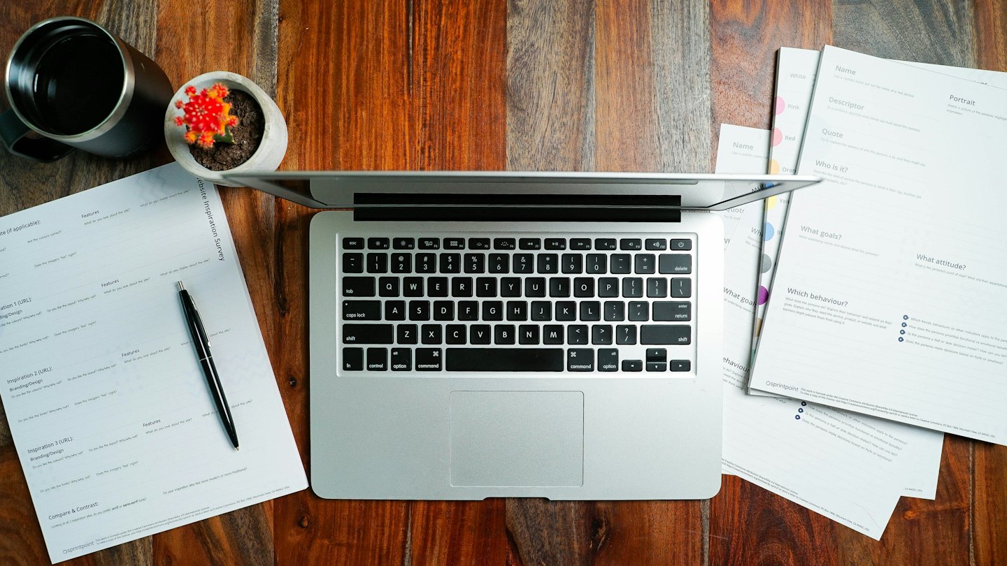 Laptop, papieren en pen op een houten bureau.